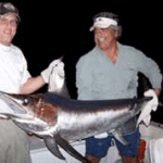 Two men holding a Marlin at night, on the back of a boat